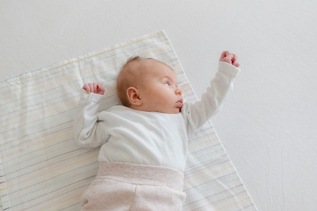 Newborn baby lying on a soft white blanket, wearing a light-colored outfit and stretching arms while looking to the side.
