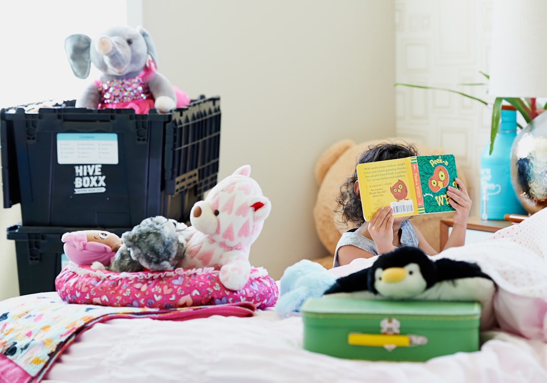 Child reading a colorful picture book on a bed surrounded by stuffed animals and toys, with moving boxes in the background.