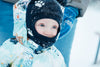 A close-up of a baby dressed in a winter snowsuit and a warm hat, with snowflakes on the hat, standing outside in the snow.