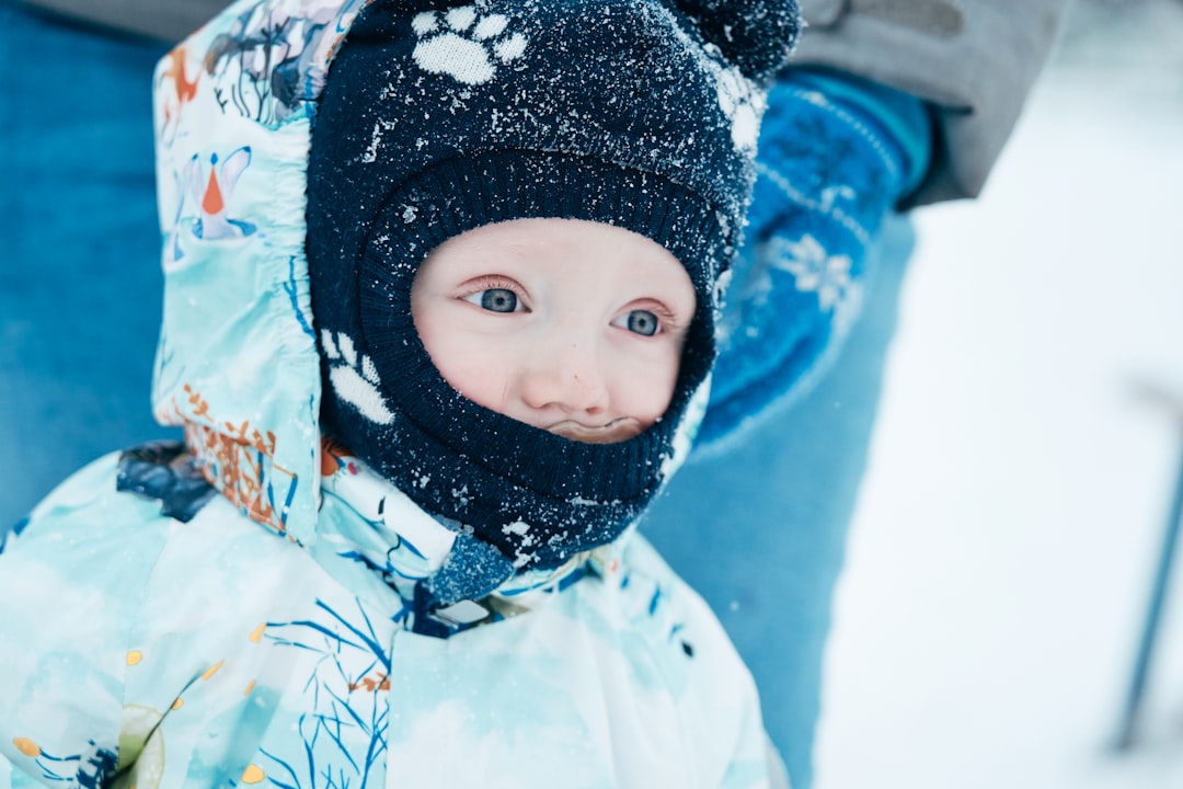 A close-up of a baby dressed in a winter snowsuit and a warm hat, with snowflakes on the hat, standing outside in the snow.