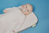 An overhead shot of a baby in a white sleepsuit lying on a quilted, white and gray mat with a green tag, sucking on a pacifier, against a solid blue background.