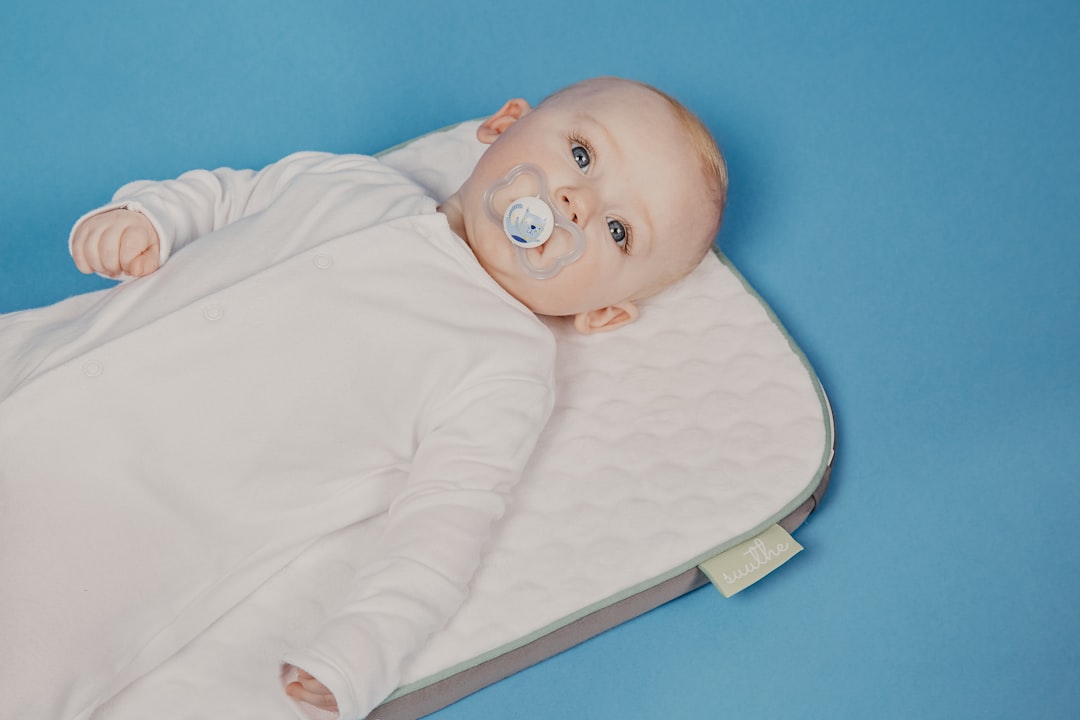 An overhead shot of a baby in a white sleepsuit lying on a quilted, white and gray mat with a green tag, sucking on a pacifier, against a solid blue background.