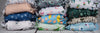 : A horizontal banner image showing three stacks of folded, colorful cloth diapers with various patterns, including koalas, flamingos, avocados, and tropical plants.