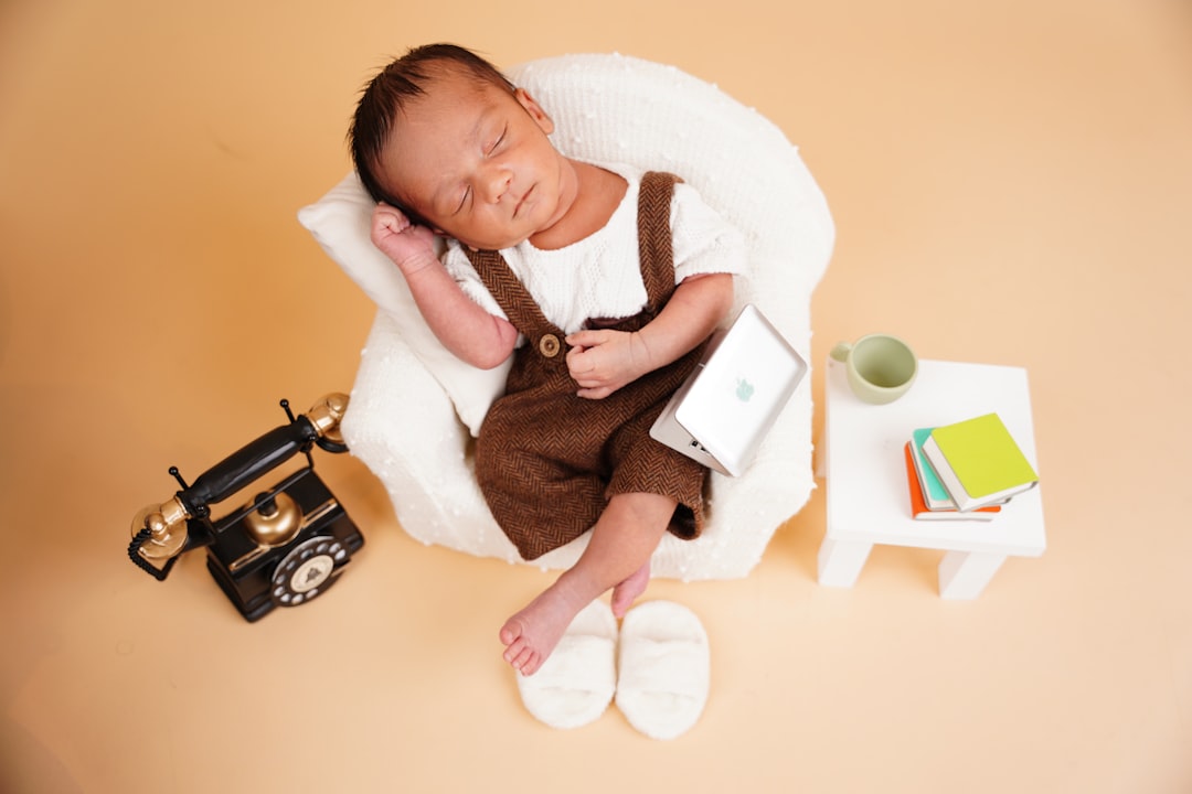 A newborn baby sleeping in a plush chair, dressed in overalls, posed with a miniature laptop, a vintage telephone, and tiny slippers for a conceptual photo shoot.
